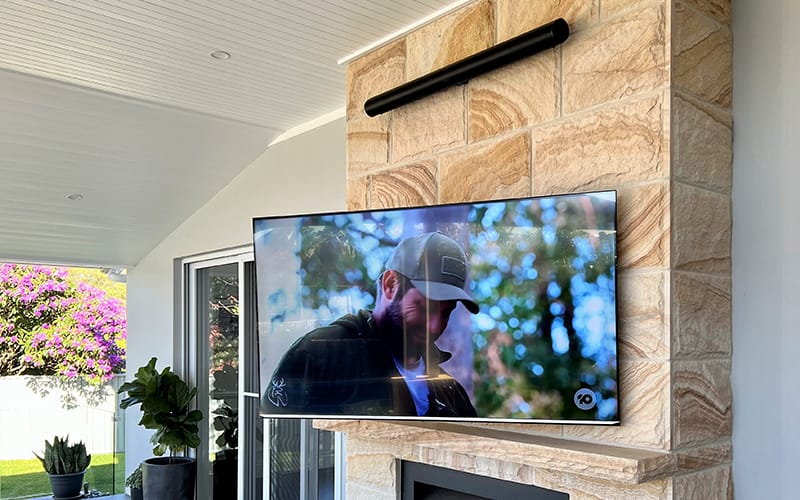 Outdoor television mounted on a sandstone wall under a covered alfresco area, with a weatherproof light bar above